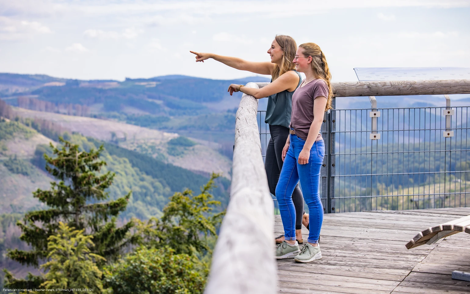 Zwei Frauen stehen auf einer hohen Brücke und blicken in die Ferne.