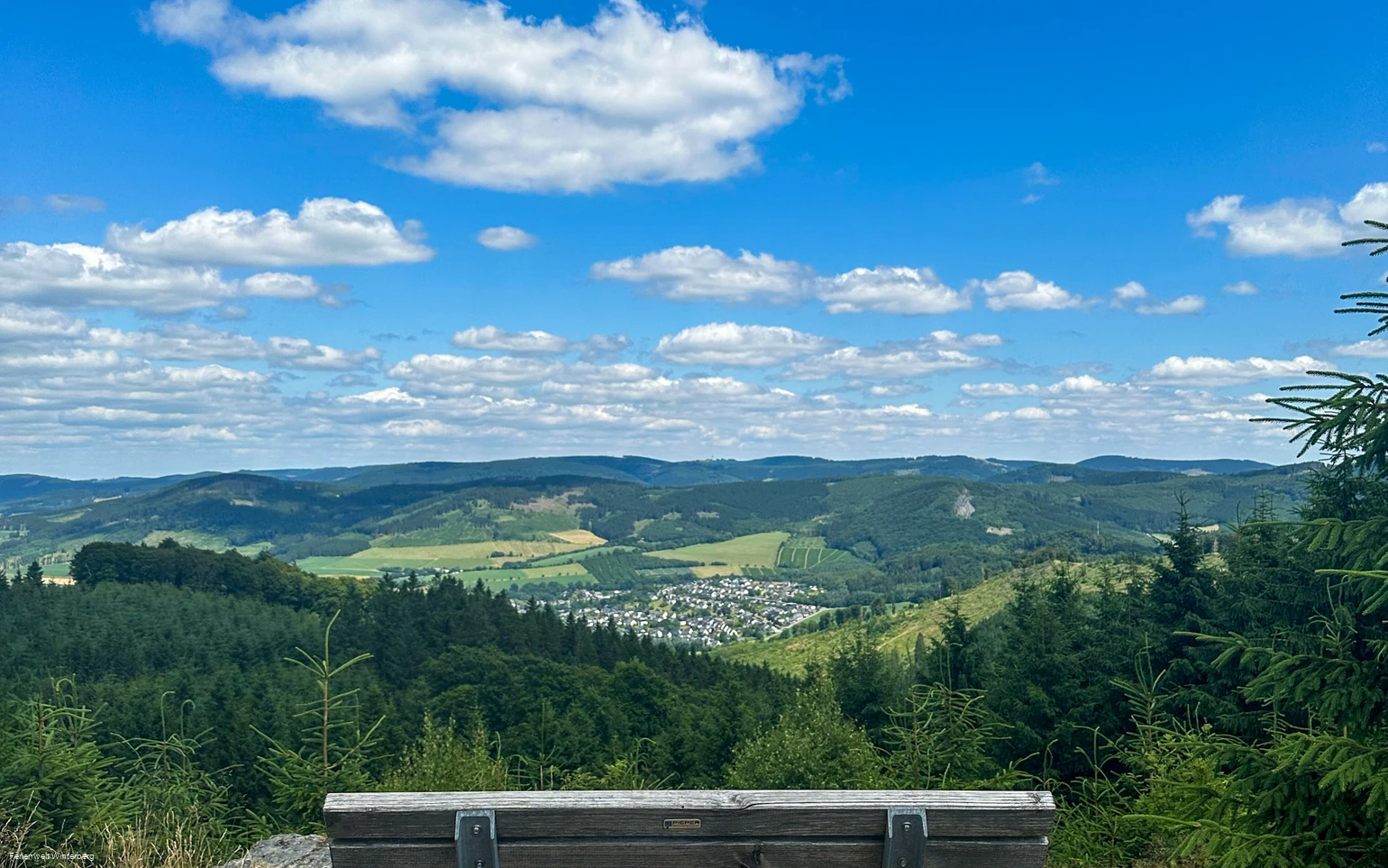 Bank auf einem Felsen mit weiter Aussicht über Wiesen und Wälder und einer Ansammlung von Häusern.