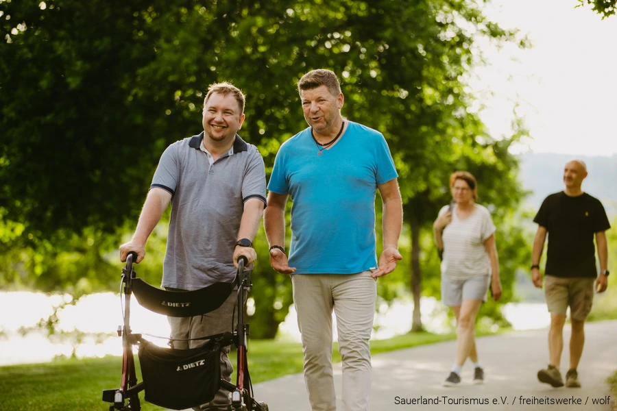 Im Vordergrund spazieren zwei M&auml;nner. Einer davon benutzt einen Rollator. Im Huntergrund spazieren noch eine Frau und ein Mann.