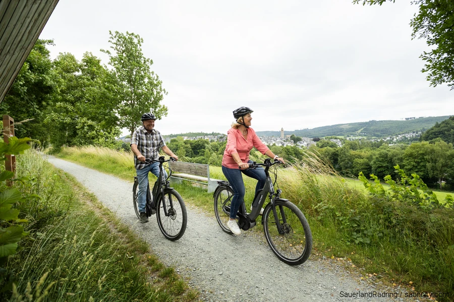 Ein &auml;lteres Paar f&auml;hrt auf E-Bikes durch die gr&uuml;ne Natur. Im Hintergrund liegt Schmallenberg.