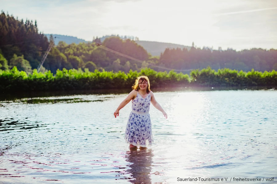 Junge Frau mit Down-Syndrom steht bis zu den Knien im Wasser eines Sees und strahlt &uuml;ber das ganze Gesicht.