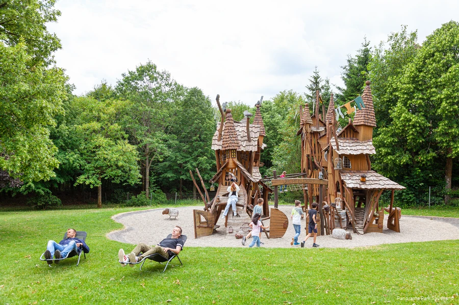 Im Hintergrund spielen Kinder auf einem Spielplatz, der aussieht wie ein Wichteldorf, und im Vordergrund entspannen zwei Väter auf Liegen.