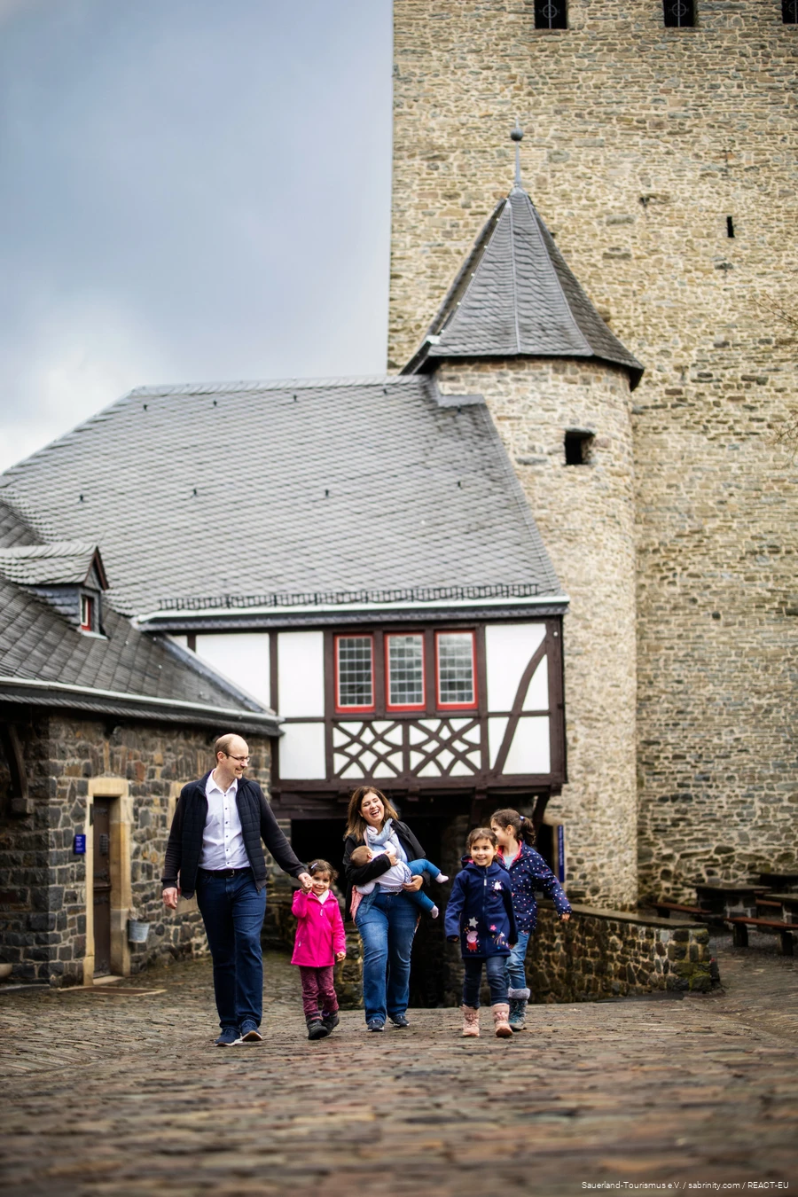 Eltern laufen mit ihren vier Kindern frühlich über den Burghof der Burg Altena mit einem Turm der Burg im Hintergrund.