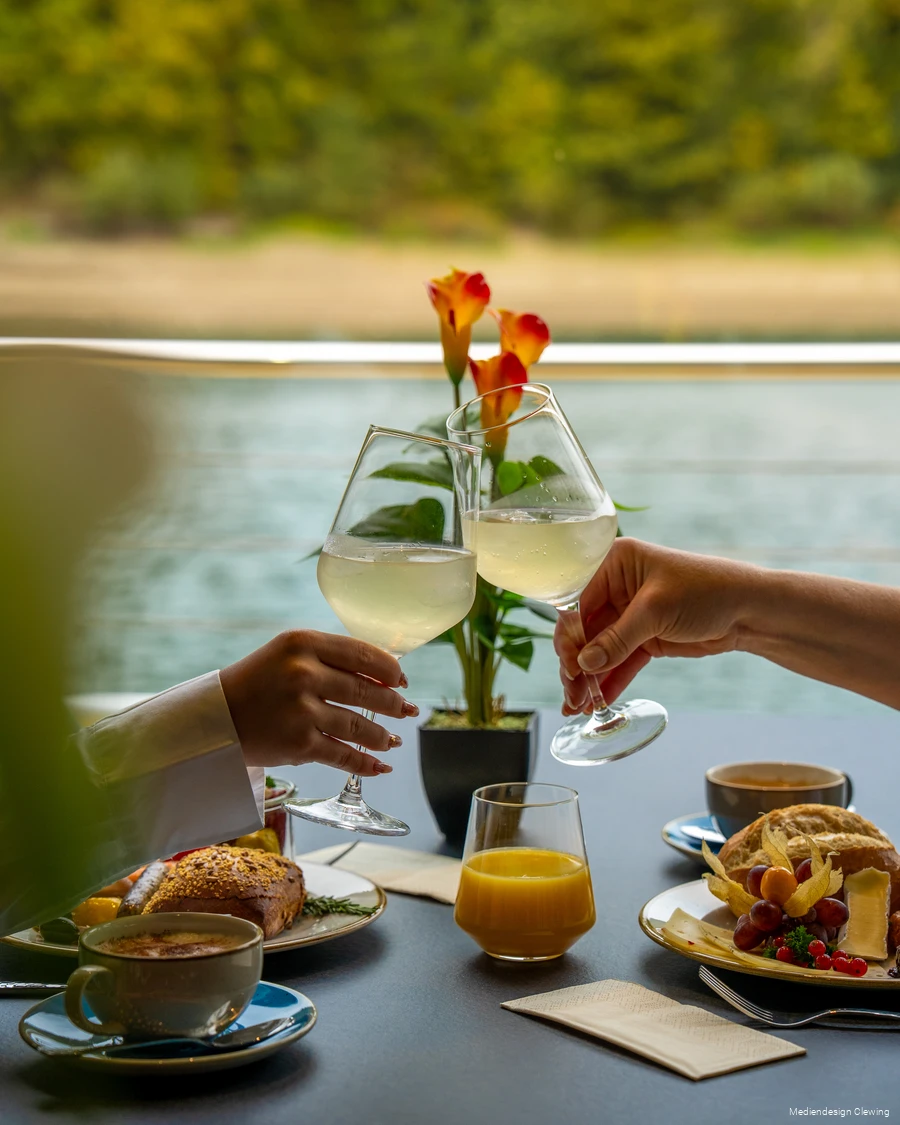 Der Tisch ist mit Frühstück gedeckt und zwei Personen stoßen mit ihren Gläsern an. Im Hintergrund sind Wasser und Ufer des Hennesees zu sehen.