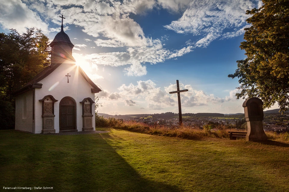 Eine Kapelle neben einem Kreuz und einer Kreuzwegstation mit blauem Himmel und der Sonne im Hintergrund.
