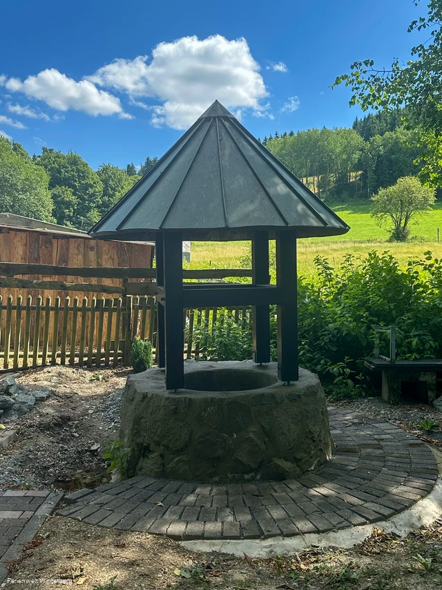 Ein überdachter Brunnen mit Wald und Wiese im Hintergrund.