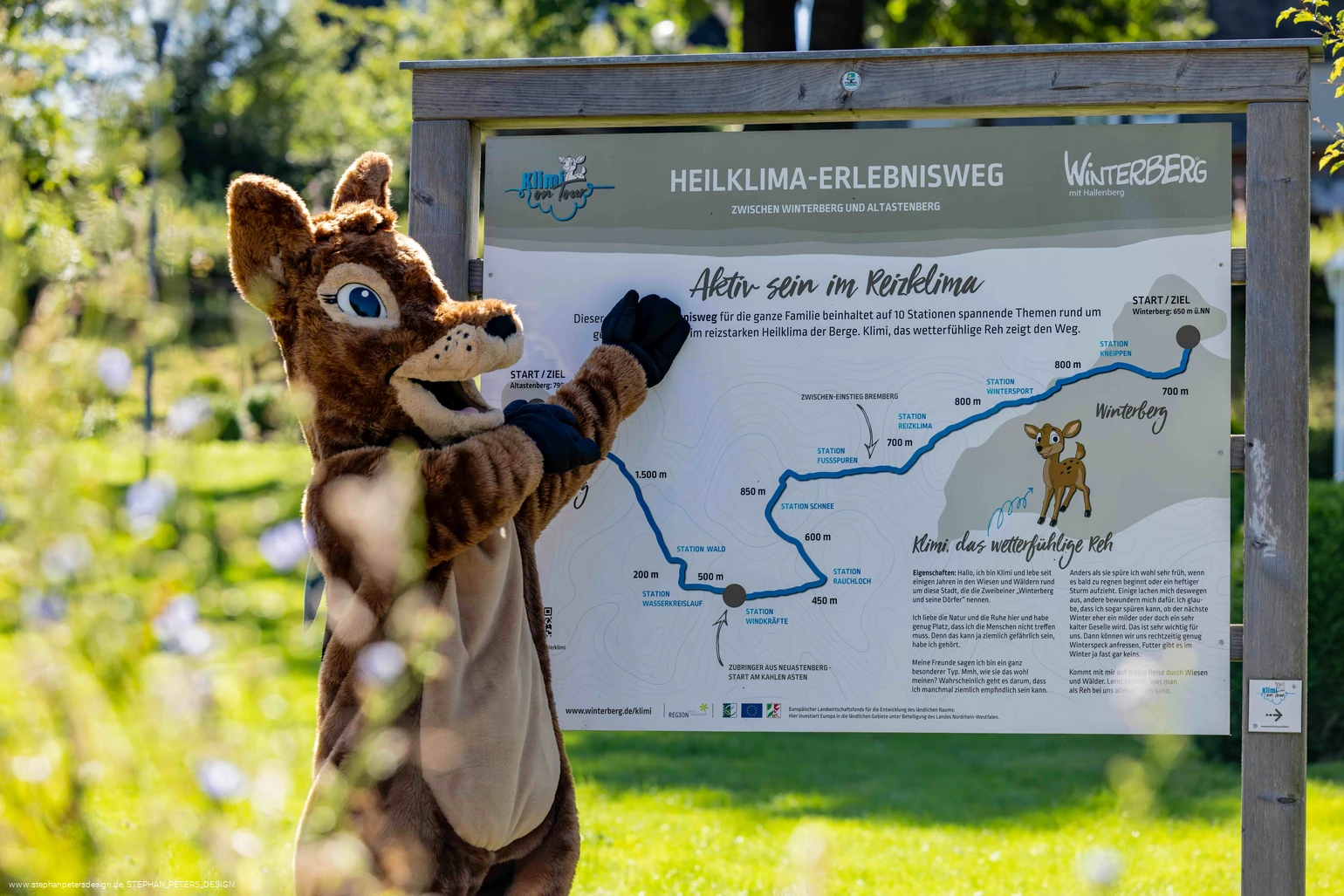 Waldi zeigt auf das Heiklima-Erlebnisweg-Schild