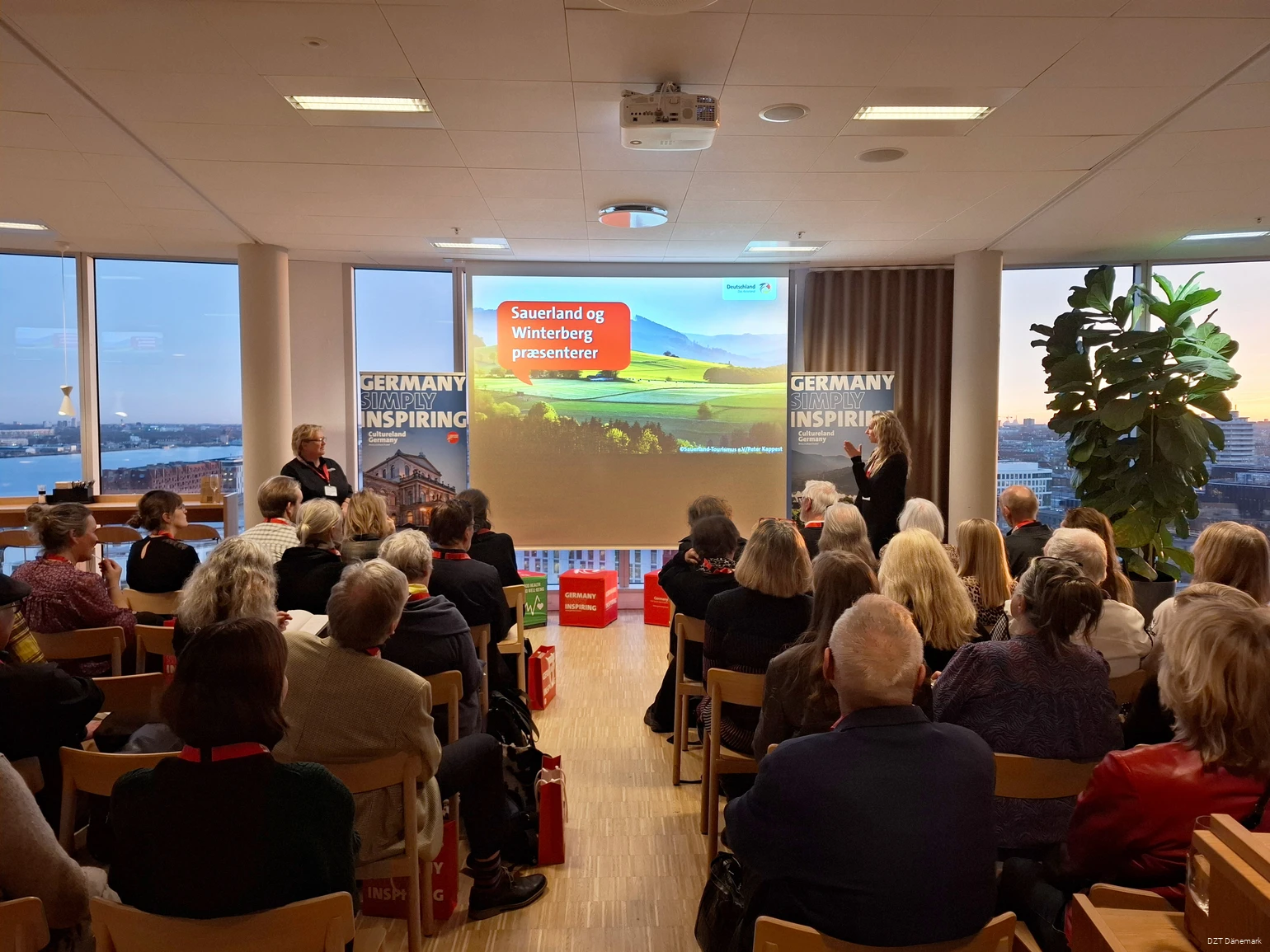 Eine größere Gruppe Menschen schaut auf eine Präsentation. Daneben stehen zwei Frauen, die vortragen.