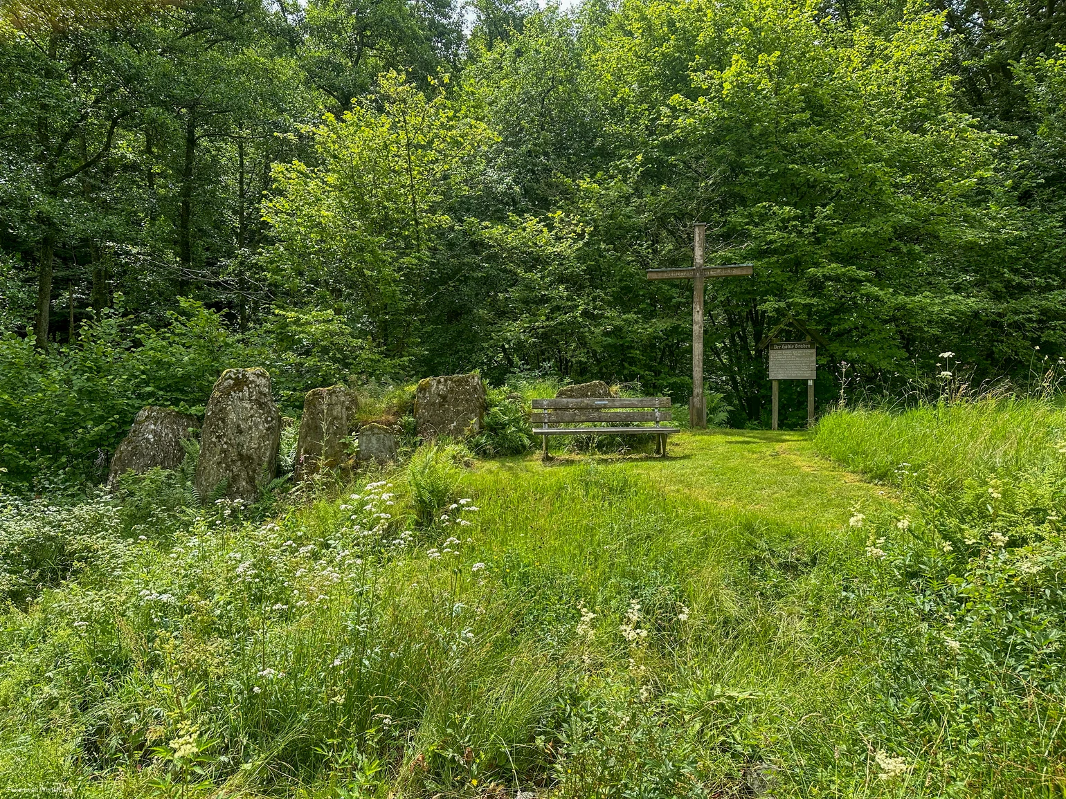 Eine Ansammlung von großen Steinen neben einer Bank und einem großen Holzkreuz, sowie einer alten hölzernen Tafel mit Informationen zu den Opfersteinen im Hohlen Graben.