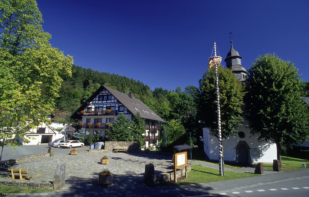 Ein gepflasterter Platz mit Sitzgelegenheiten und Informationstafeln neben einer Kapelle.