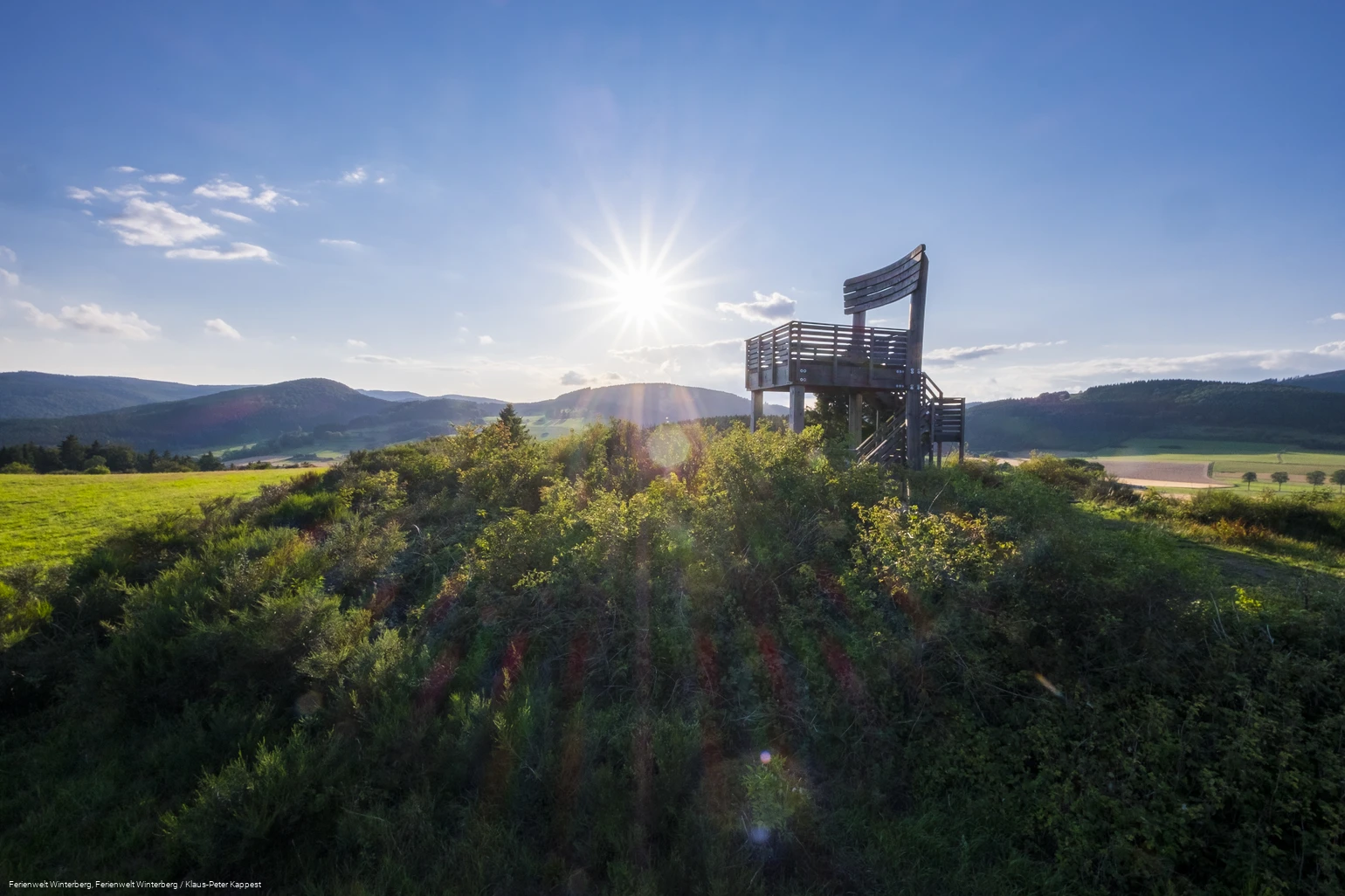 Ein Aussichtsturm in Form eines großen Holzstuhles auf einem bewaldeten Hügel vor blauem Himmel und Sonne inmitten von Wiesen und Wäldern.