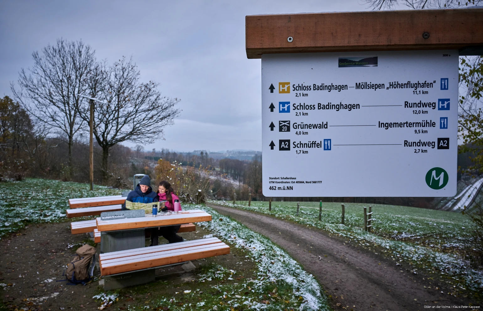 Im Vordergrund ist ein Wegweiser, im Hintergrund sitzt ein Paar auf einer Bank und betrachtet eine Karte. Es liegt eine leichte Schneeschicht.