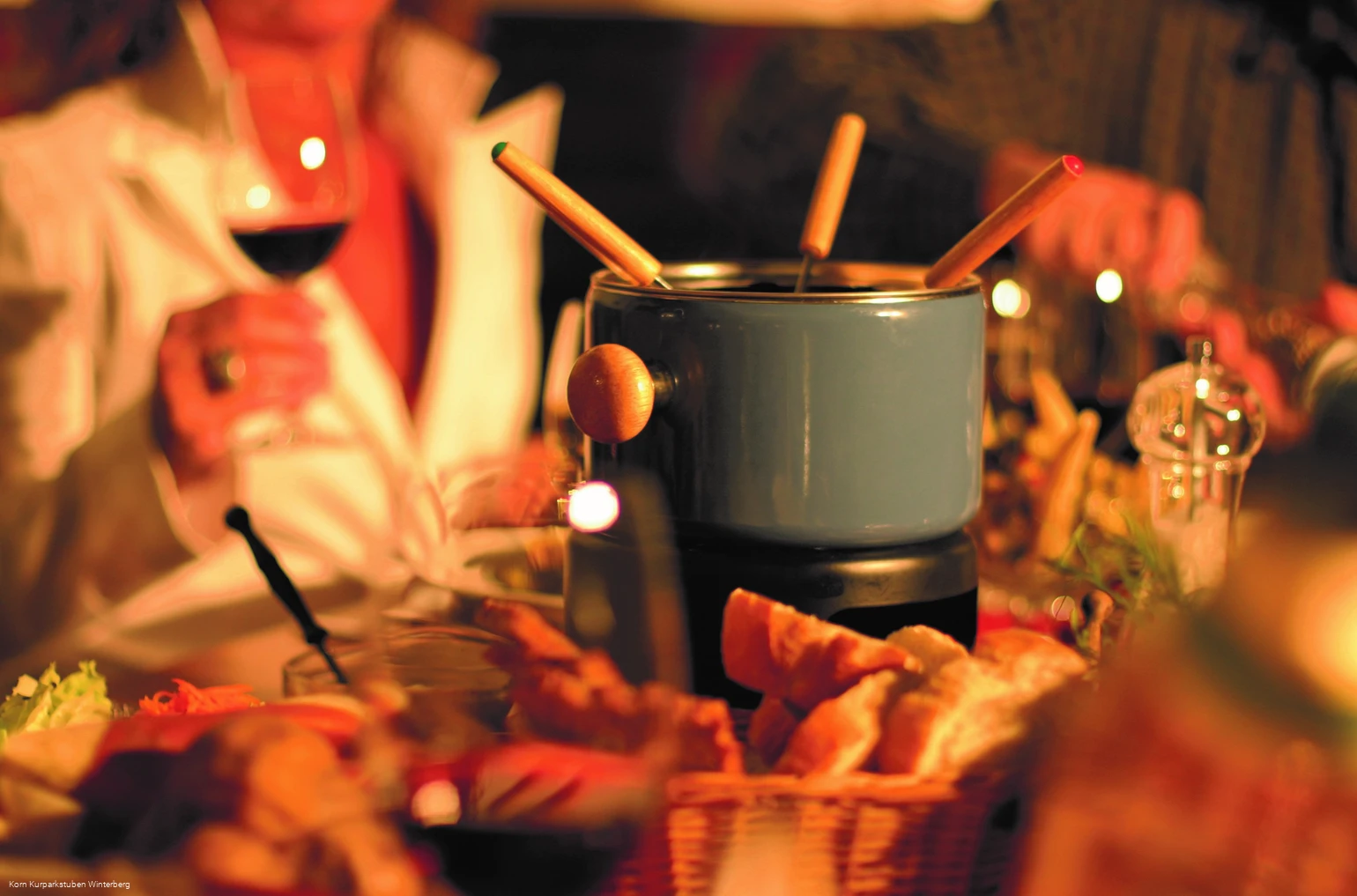 Ein Fonduetopf mit Gabeln auf einem gedeckten Tisch sowie eine verschwommene Person  im Hintergrund, welche ein Weinglas in der Hand hält.