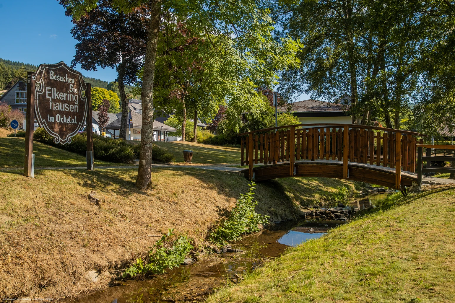 Eine hölzerne Brücke über einem Bach umgeben von Wiese und Bäumen und einem Holzschild mit der Aufschrift Besuchen Sie Elkeringhausen im Orketal.
