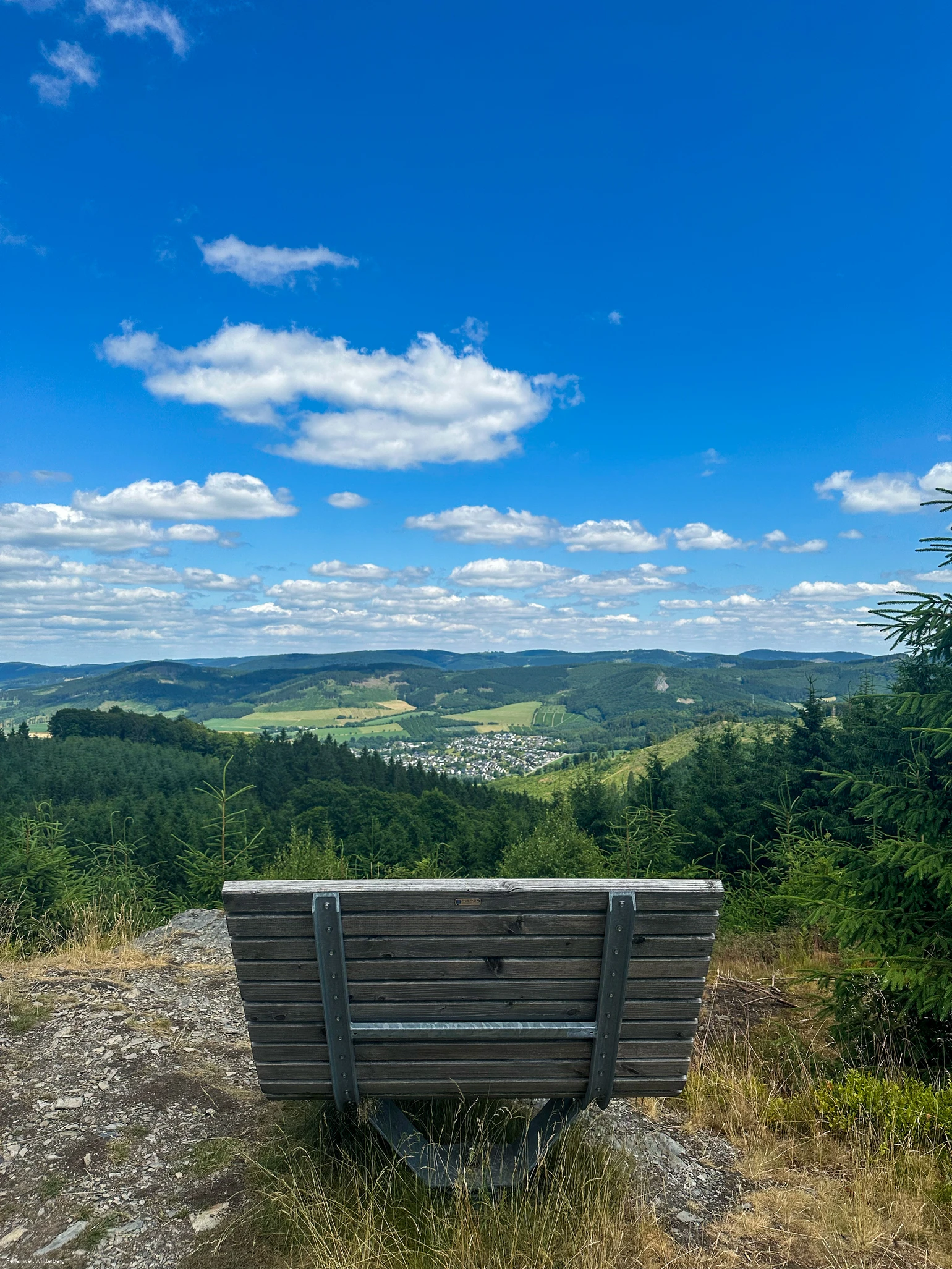 Bank auf einem Felsen mit weiter Aussicht über Wiesen und Wälder und einer Ansammlung von Häusern.