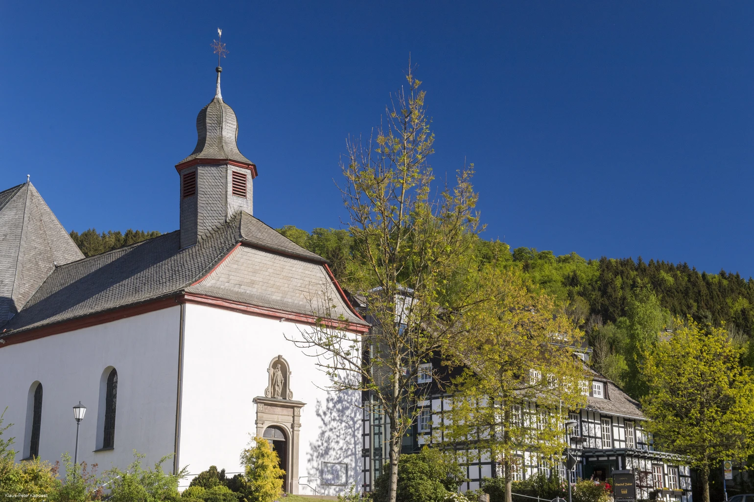 Eine Kirche vor blauem Himmel neben einem Fachwerkhaus, umgeben von Bäumen.