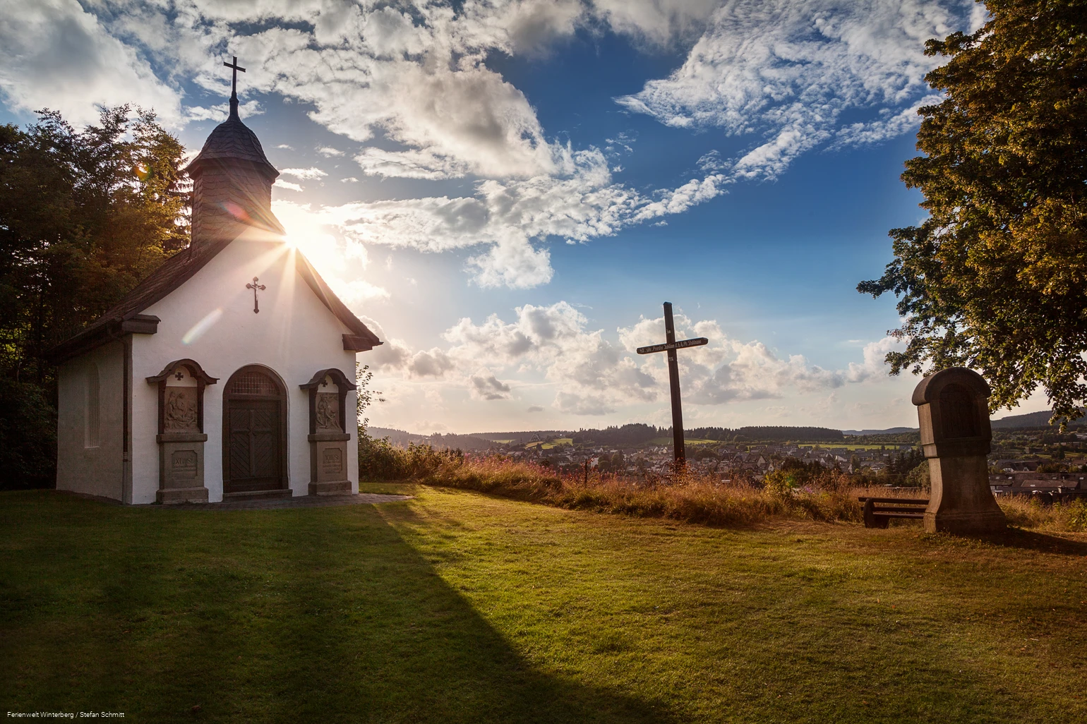 Eine Kapelle neben einem Kreuz und einer Kreuzwegstation mit blauem Himmel und der Sonne im Hintergrund.
