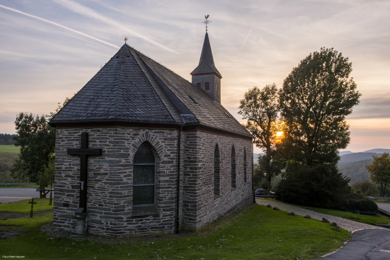 Eine steinerne Kapelle umgeben von einer grünen Wioese mit der untergehenden Sonne im Hintergrund.
